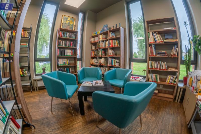 pomieszczenie z podłużnymi oknami, regałami z książkami, na środku stolik z czterema niebieskimi fotelami