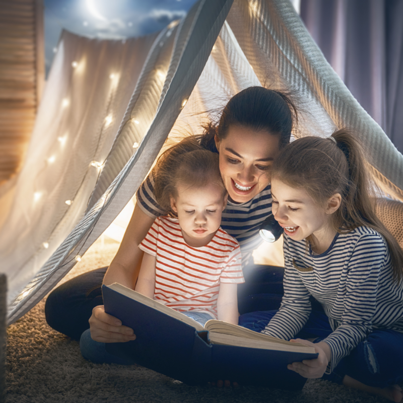 kobieta czytająca dwóm dziewczynkom przy świetle latarki
