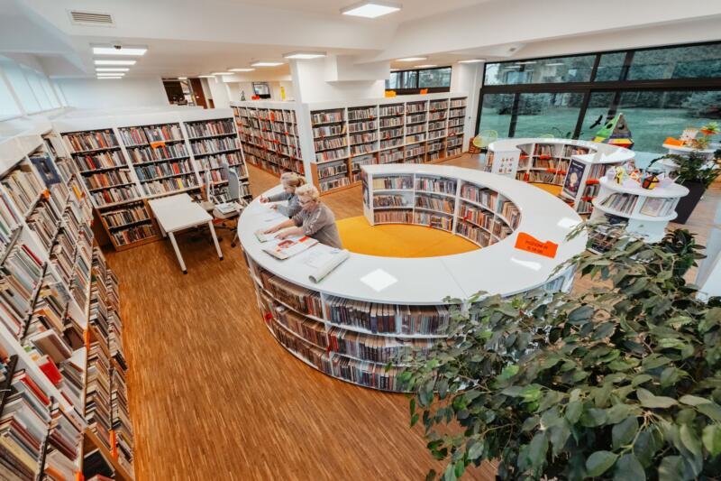 pomieszczenie biblioteki z wieloma regałami i książkami, na środku okrągły regał, przy którym stoją dwie kobiety