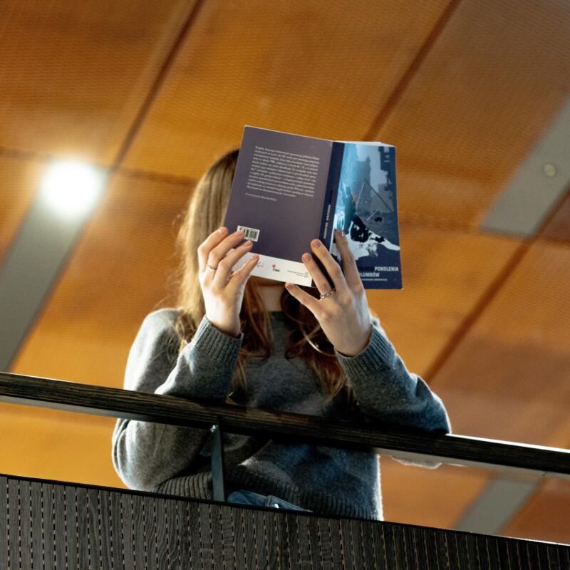 kobieta stoi na balkonie Biblioteki Narodowej, czyta książkę, która zasłania jej twarz